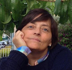 Woman resting her chin on her hand outdoors, with green leaves in the background and a gentle smile.