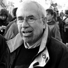Older man with glasses in a crowded outdoor event, smiling in a black-and-white photo.