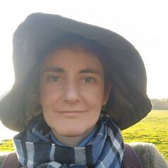 Portrait of a woman in a wide-brim hat and striped scarf, standing in a sunlit grassy field and looking at the camera.