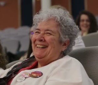 Smiling elderly woman with gray curly hair, wearing a white jacket and buttons, seated in a conference setting.