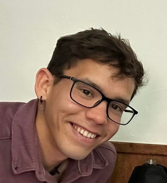 Smiling person with short dark hair and black rectangular glasses, wearing a purple shirt indoors.