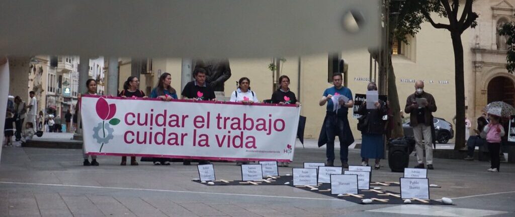 Group of protesters holding a banner reading 'cuidar el trabajo cuidar la vida' in a plaza/ street scene with onlookers and signs on the ground behind them