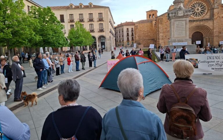 Crowd gathered in a town square around a blue and red camping tent near a church and historic buildings.