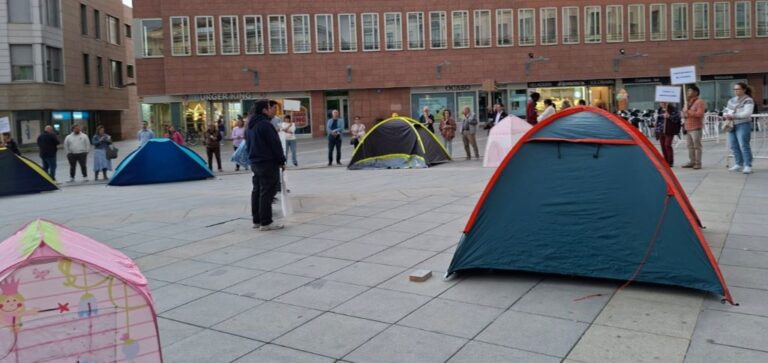 Tents pitched in a city plaza with people standing around and storefronts in the background, suggesting a public gathering or demonstration.