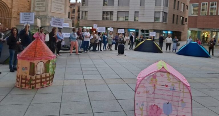 Crowd of people holding protest signs in a city square with colorful tents in the foreground.