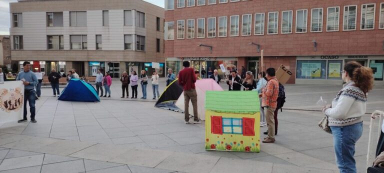 People gathered in a plaza with small tents and a colorful cardboard house prop in the foreground, suggesting a community or protest event earlier in the day at a shopping district.