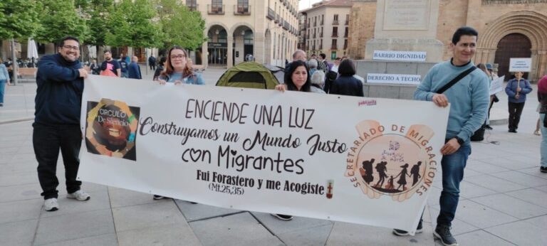 Group of people standing in a plaza holding a banner that reads: 'Encende una luz Construyamos un Mundo Justo con Migrantes' with logos on the sides and a circular emblem about migration rights in the background.