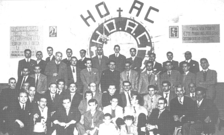 Large group of men in suits posing for a formal group photo in front of a wall with a circular emblem on it.