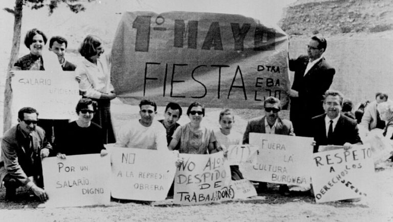 Black-and-white photo of a May Day rally: a group of workers posing with a large banner reading '1º MAYO FIESTA' and signs demanding a decent salary, no worker layoffs, end repression, and respect for workers.