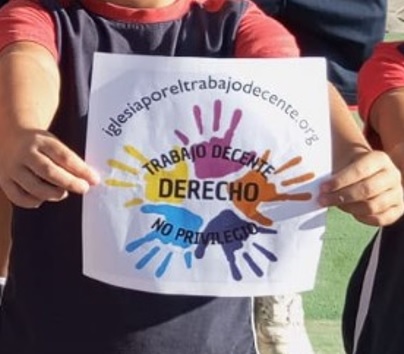 Person holding a sign with rainbow handprints and the Spanish phrases 'Trabajo Decente', 'Derecho', 'No Privilegio'.