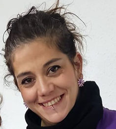 Smiling woman with dark hair tied back, wearing a black scarf and purple top, with pink flower-shaped earrings.