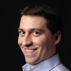 Smiling man with short dark hair wearing a light blue shirt, head-and-shoulders portrait against a dark background.