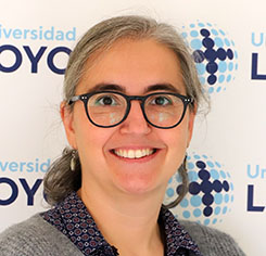 Portrait of a smiling woman wearing dark-framed glasses, in front of a backdrop with university logos.