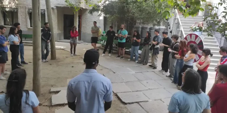 Group of people standing in a loose circle outdoors, listening to someone in the center.