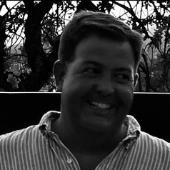 Smiling man wearing a striped shirt outdoors with trees in the background.