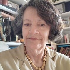 Portrait of a smiling woman with short gray hair, wearing a beige blazer and wooden bead necklace, in a bookshelf-filled room on a home office backdrop.