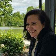 Smiling woman in a dark blazer sits outdoors by a lake with trees in the background.
