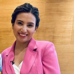 Smiling woman with short dark hair wearing a pink blazer, standing against a wooden wall background.