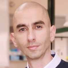 Close-up portrait of a bald man with light stubble, wearing a white shirt and looking at the camera.