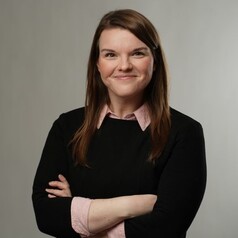 Portrait of a smiling woman with brown hair, arms crossed, wearing a black sweater over a pink collared shirt, against a light gray backdrop.