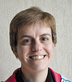 Person with short brown hair and a warm smile, wearing a red jacket against a light textured wall.