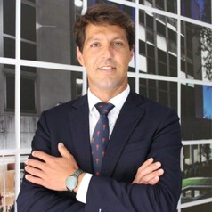 Businessman in a dark suit with a red tie, arms crossed, standing in front of a glass office building.
