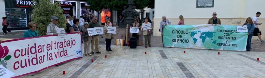 Group of people in a plaza holding banners about workers' rights and solidarity with migrants, with red candles lined along the ground.