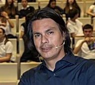 Portrait of a man with shoulder-length dark hair in a blue shirt, speaking at a conference with an audience in the background, focused expression.