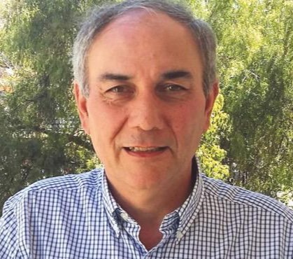Portrait of a middle-aged man with gray hair wearing a blue-and-white checkered shirt, outdoors with trees in the background.