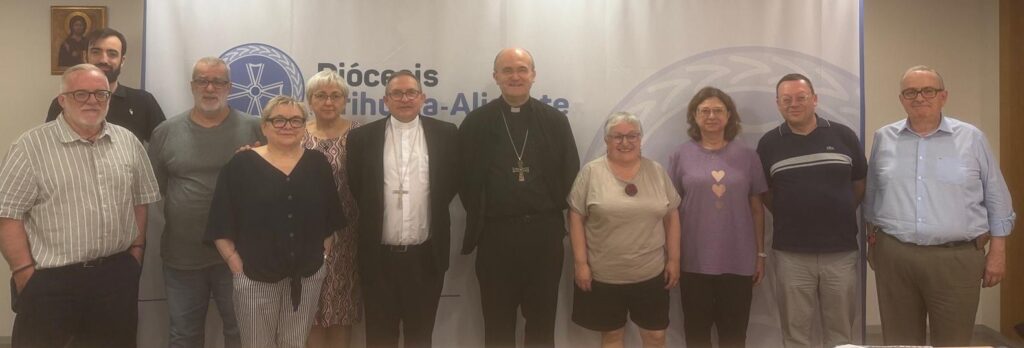 Group photo of twelve people, including two clergy in clerical collars, standing in front of a banner at a church-related event.