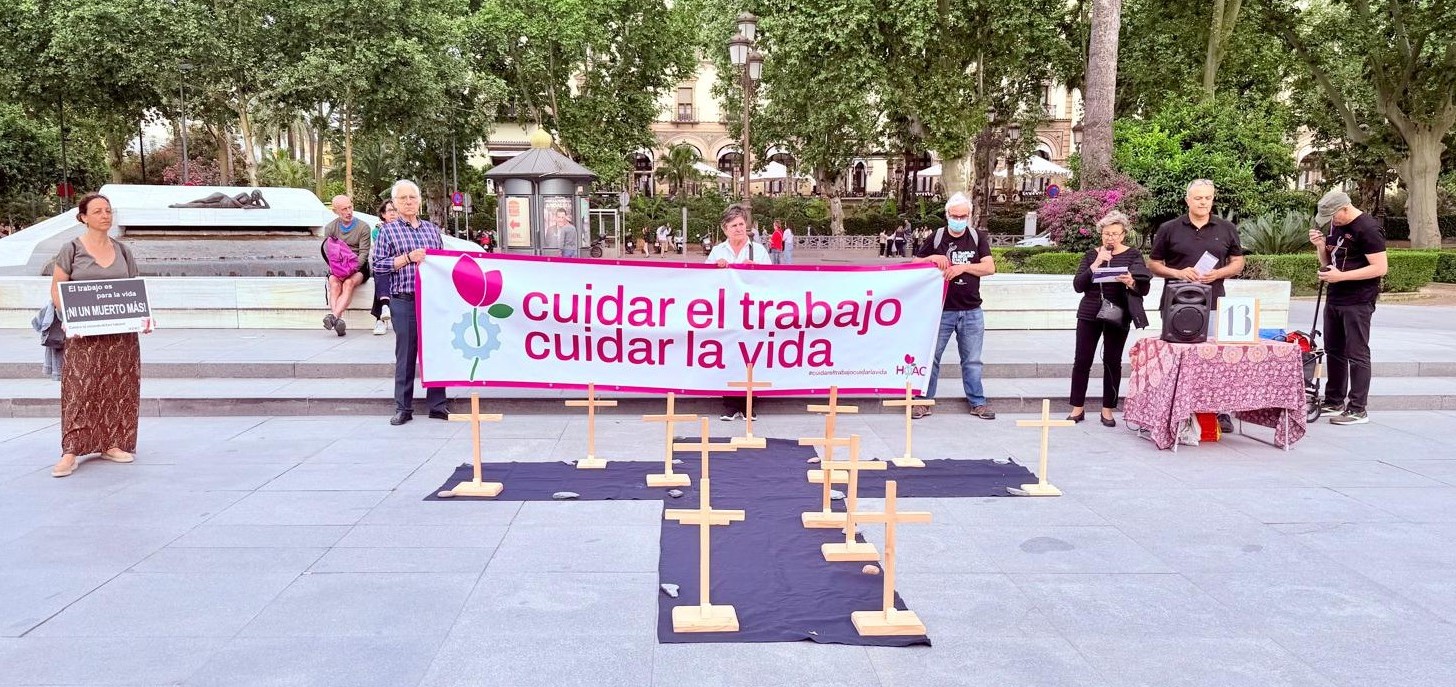 Group of protesters standing behind a pink-and-white banner that reads 'cuidar el trabajo cuidar la vida' in a plaza, with wooden crosses arranged on dark fabric on the ground as a memorial display.