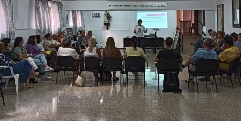 Adults seated in a semicircle in a bright room, listening to a speaker at the front with a projector display.