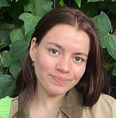 Close-up portrait of a woman with brown hair wearing a tan jacket, smiling, with green leafy background.