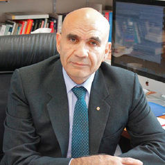 Businessman in a dark suit and teal tie sits at a desk in an office, facing the camera with bookshelves and a computer monitor in the background.