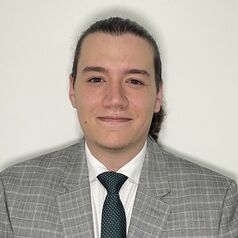 Portrait of a man with dark hair tied back, wearing a gray checkered suit and teal tie, with a neutral expression against a white background.