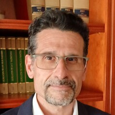Portrait of a man with short dark hair and glasses, wearing a navy blazer, in front of a wooden bookshelf.