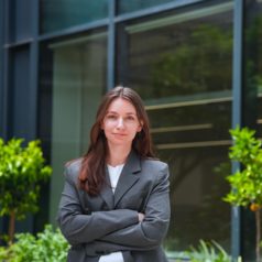 Professional woman with brown hair in a gray blazer standing outside a glass-front office building, arms crossed.