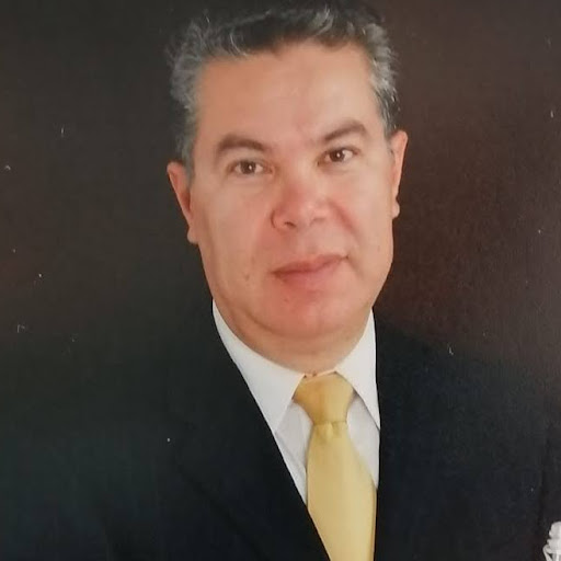 Portrait of a man in a black suit with a pale yellow tie, facing the camera against a dark background.