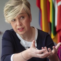 Middle-aged woman speaking and gesturing with hands during a discussion, colorful flags in the background.