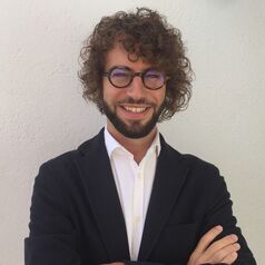 Smiling man with curly hair and glasses, wearing a dark blazer and white shirt, arms crossed against a light wall.