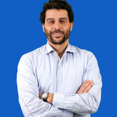 Man with arms crossed, smiling, wearing a light-blue shirt against a solid blue background.