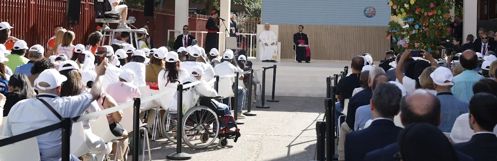 Pope Francis speaks from a stage in an outdoor ceremony, with a large seated audience and photographers on the left.
