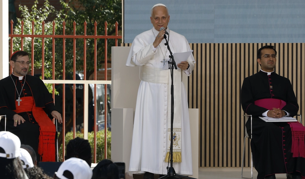 A Catholic cleric in white vestments speaks at a microphone, flanked by two other clergy in dark robes at an outdoor event behind a wooden fence.