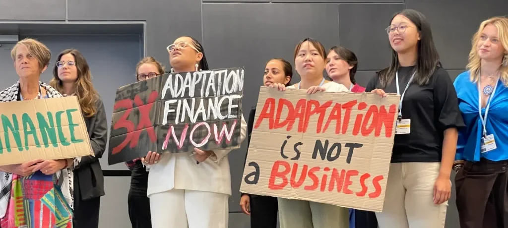 Diverse group of people indoors holding handmade protest signs about climate finance, including 'Adaptation Finance Now' and 'Adaptation is Not a Business'.