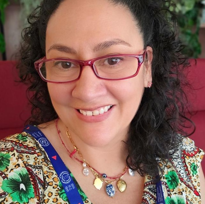 Smiling woman with red glasses and curly dark hair in a colorful patterned top, wearing layered necklaces and a blue lanyard against a pink background.