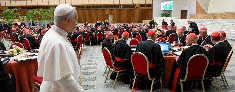 A white-robed religious leader walks past a banquet-style hall filled with cardinals in black robes with red sashes at round tables, under a stained glass dome.