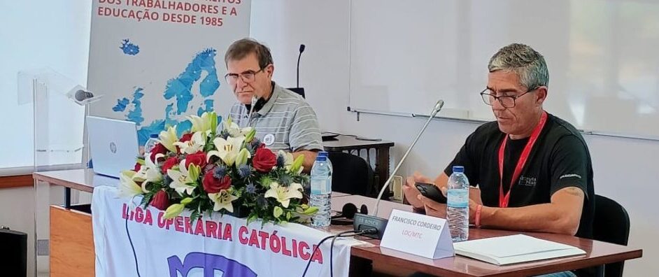 Two men sit at a panel desk with laptops, microphones, and water bottles; a floral centerpiece and banner with a globe emblem sit in front of them.