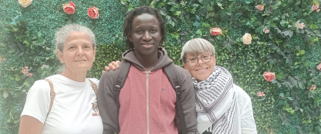 Three people posing together in front of a green floral wall: an older woman on the left, a young man center, and an older woman on the right, all smiling.