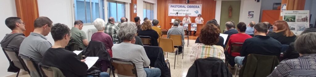 Community meeting in a hall with a panel at the front and an audience of adults listening.