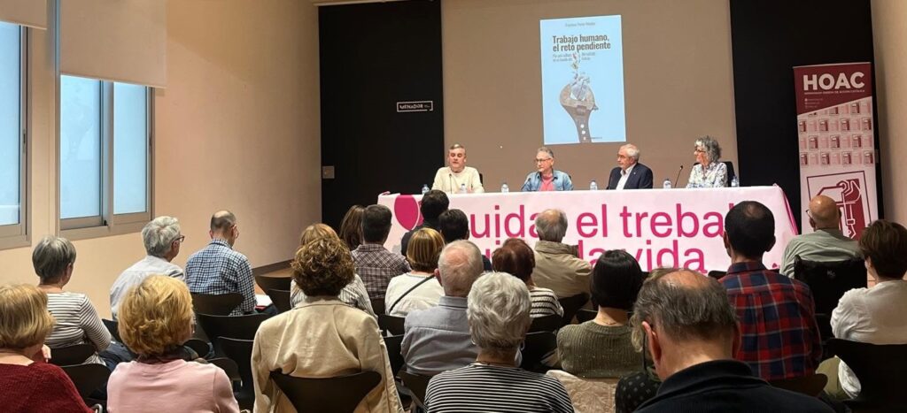 Panel discussion at a conference: five speakers sit behind a pink bannered table while an audience listens, with a poster on the screen reading 'Trabajo humano, el reto pendiente'.